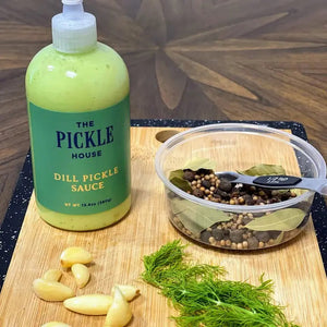 A green bottle labeled "PICKLE HOUSE - DILL PICKLE SAUCE" with a white nozzle is showcased on a plain, light gray background. The versatile 9.5 oz (269g) sauce by THE PICKLE HOUSE can be used as a salad dressing, dipping sauce, or marinade.