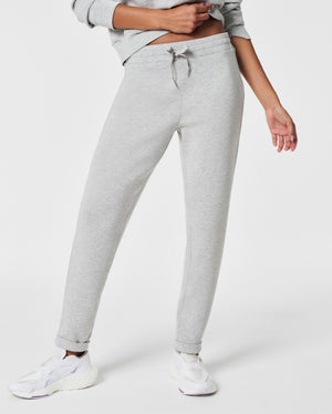 A person stands against a white background, smiling and looking down with one hand touching their hair. They are wearing a matching light beige SPANX AIRESSENTIALS TAPERED PANT and sweatshirt set made from ultra drapey, lightweight spacer fabric, paired with white sneakers.