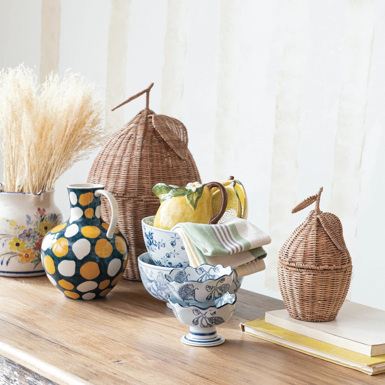 A wooden table adorned with various decorative items, including a vase with dried grass, a colorful polka-dotted pitcher, a Hand Stamped Footed Bowl with Scalloped Edges by Creative Coop featuring intricate blue botanical prints, a yellow pitcher with limes, neatly folded towels, and wicker baskets shaped like huts.