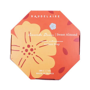 A round, wrapped bar of BAUDELAIRE - SWEET ALMOND 7OZ BOXED BATH BAR with a weight of 200 grams (7 ounces). The label features text in both French and English, along with an orange flower graphic. Made in France and enriched with shea butter, it is imported by Baudelaire, Swanzey, NH.