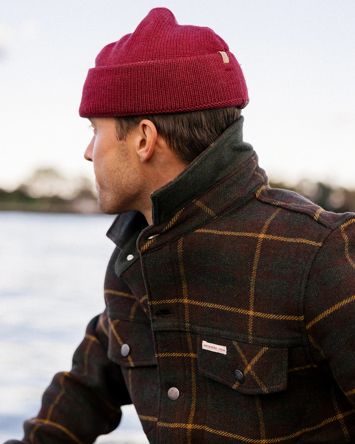 The AMUNDSEN SPORTS Wool Jacket is a unisex dark plaid overshirt with a brown and mustard check pattern, long sleeves, buttoned cuffs, and a small red, white, and blue striped tag near the back collar. Laid flat, viewed from behind.