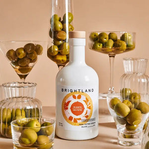 A white glass bottle with a wooden cap labeled "Brightland" and "100% Castelvetrano Olive Oil" in black text. The bottle boasts colorful abstract artwork of hands holding a garlic bulb, capturing the essence of Sicily. This single estate extra virgin olive oil has a net weight of 375 ml / 12.7 fl oz.