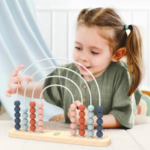 A young child with pigtails is playing with the SPEEDY MONKEY - RAINBOW ABACUS. The abacus, an early educational preschool toy from the SPEEDY MONKEY brand, is made from sustainably sourced wood and features five arcs adorned with colorful beads in shades of pink, blue, and gray. The child, wearing a green shirt and seated indoors against a blurred background, is focused and engaged.