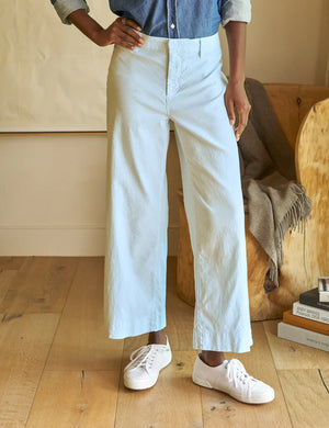 A person in a denim shirt and FRANK AND EILEEN - WEXFORD WIDE LEG LINEN PANT with white sneakers stands with their back to the camera. Nearby, a wooden chair holds a blanket, and books are stacked on the floor.