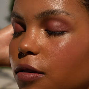 A close-up of a person with curly dark hair, a serious expression, and warm makeup featuring the universally flattering, weightless JASON WU - HOT FLUFF LIP/EYE/CHEEK by JASON WU BEAUTY on their face. They are looking directly at the camera with a neutral background.