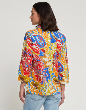 A woman with shoulder-length dark hair is shown from behind, wearing the HINSON WU Aileen 3/4 Sleeve Paisley Print Top, which features a colorful blend of red, yellow, blue, and orange shades. She has paired it with light blue denim jeans against a plain white background. This best-selling top from HINSON WU exudes style and vibrancy.