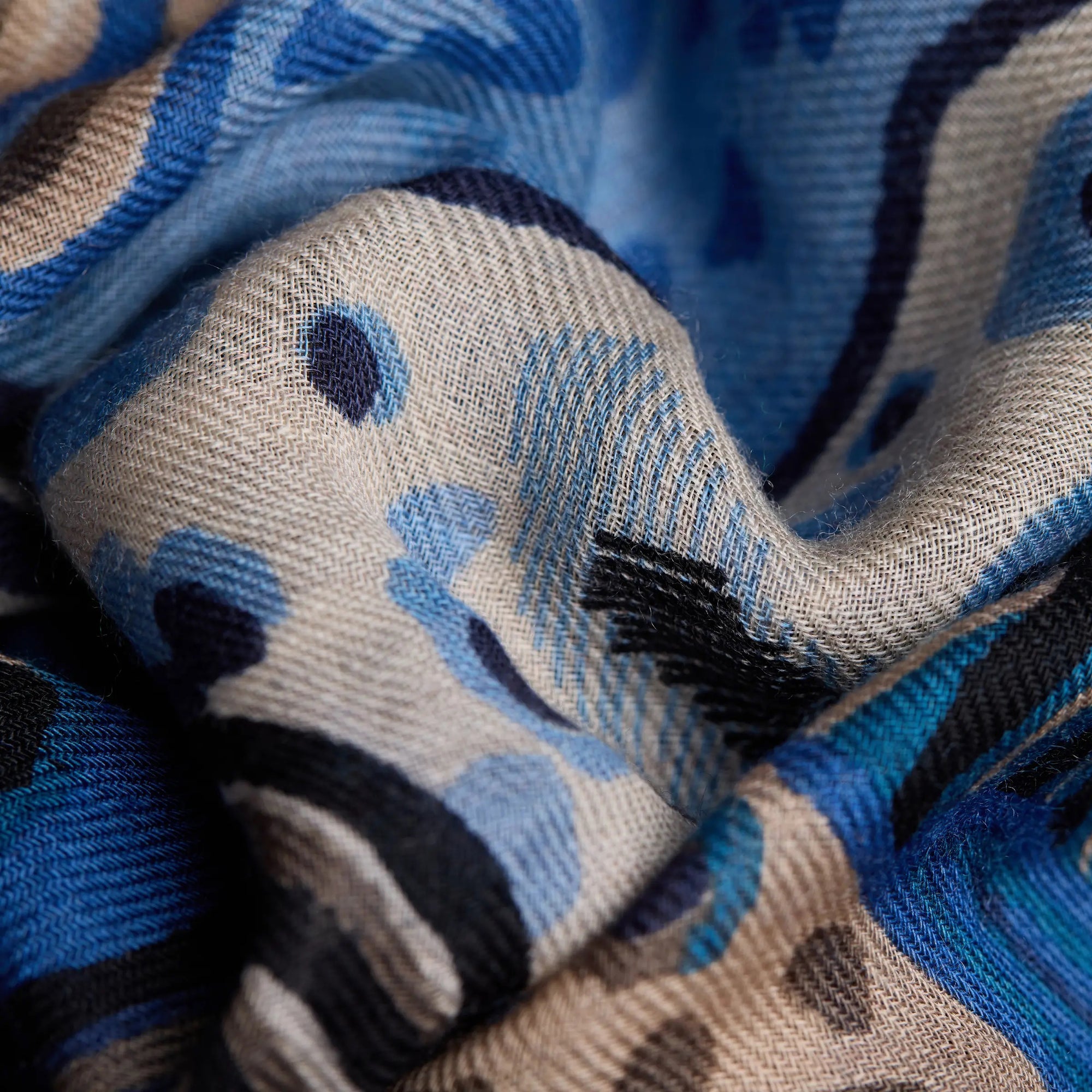 Close-up of an INOUI EDITIONS - WESTERN SQUARE SCARF featuring a blue and beige abstract pattern. The 100% wool fabric appears soft and slightly wavy, showcasing intricate weaving and a smooth texture. The unique design includes shades of blue, black, and beige with curved and dotted shapes. Perfect for adding a stylish touch to any outfit.