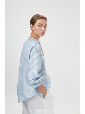 A person with light brown hair pulled back is wearing an oversized LANHTROPY - SAKE LINEN SHIRT by LANHTROPY, made of organic linen fabric, featuring a light blue and white striped pattern with buttons down the front and a mandarin collar. The sleeves are rolled up, and they have their left hand in the pocket of their white pants. The background is plain white.