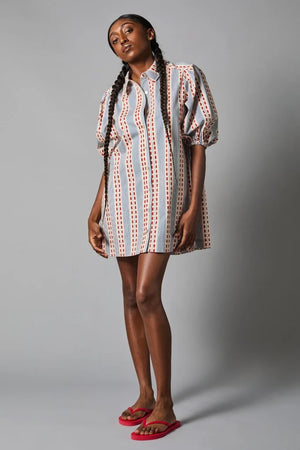 A person stands with their back to the camera, wearing the eco-friendly HOPE FOR FLOWERS TUCKED SLEEVE SHIRTDRESS, GOTS Certified with vertical stripes and dots in red, white, and blue. The dress has puffed sleeves and pairs perfectly with red open-toe sandals. The background is gray.