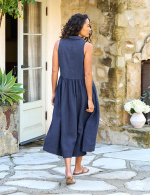 The FRANK & EILEEN - Daphne Sleeveless Popover Dress is a sleeveless, dark blue midi dress with a fitted bodice, collared neckline, and a lightly pleated, flowing skirt. Crafted from washed linen, the dress also features handy side pockets. Displayed on a white background.