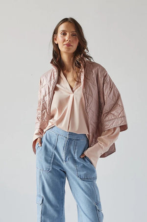 A person with shoulder-length hair stands facing sideways, wearing the ADROIT ATELIER - RUBY QUILTED CAPE, a light pink, short-sleeved jacket with a 100% Nylon shell, paired with light blue jeans. The background is plain white. The individual gazes off to the side, showcasing the back and side profile of the outfit from ADROIT ATELIER.