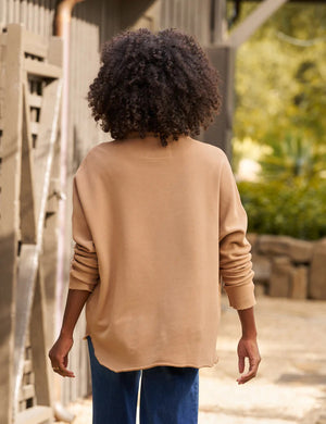 A person with curly hair stands outside in front of a building, wearing FRANK & EILEEN's Effie Long Sleeve Funnel Neck Capelet paired with blue jeans. They have a relaxed pose, accentuated by the soft drape of the capelet. The background features a wooden structure and lush greenery.