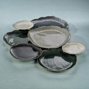 The image showcases the ZODAX ENAMELED 7 CLUSTER SERVEWARE SET featuring ceramic plates in diverse sizes and shades of black, grey, and white. Each plate boasts a slightly uneven, textured surface with irregular edges that lend an artisanal aesthetic. Arranged overlapping one another, the complete setup measures 72.39 cm x 48.26 cm x 3.81 cm (28.5 in x 19 in x 1).