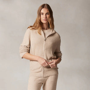 A person with long hair, wearing Kashwere's KASHWERE - RELAXED HOODIE made from Kash-ease yarn, has their hands in the front pockets of a light beige zip-up. The hood is up against a neutral, textured background. This wardrobe staple from KASHWERE looks both comfortable and casual.