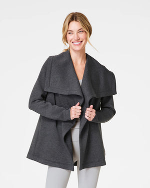 A person with long hair tied back is wearing the SPANX AireEssentials Drapefront Cardigan, crafted in super-soft spacer fabric. They are standing and facing away from the camera against a plain white background, showcasing the essence of everyday layering.