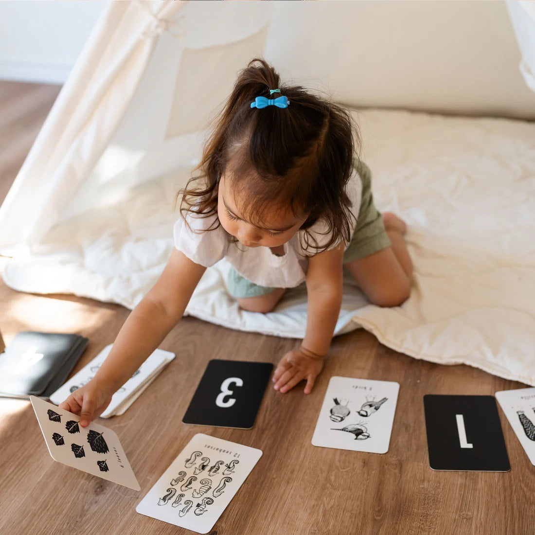 A woman and a young child sit together on the floor inside a white play tent. The woman is holding one of Wee Gallery&#39;s Nature Number Cards and showing it to the child. In front of them, four black cards with the numbers 1 to 4 from Wee Gallery&#39;s Nature Number Cards set are lined up on the floor.