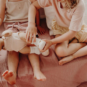 A close-up of a baby wearing OLLI ELLA USA DINKUM DOLL SHOES in Mallow Pink and white socks. The baby is seated on a soft, pale pink textured blanket, dressed in light brown clothing. Only the lower half of the baby's body is visible.