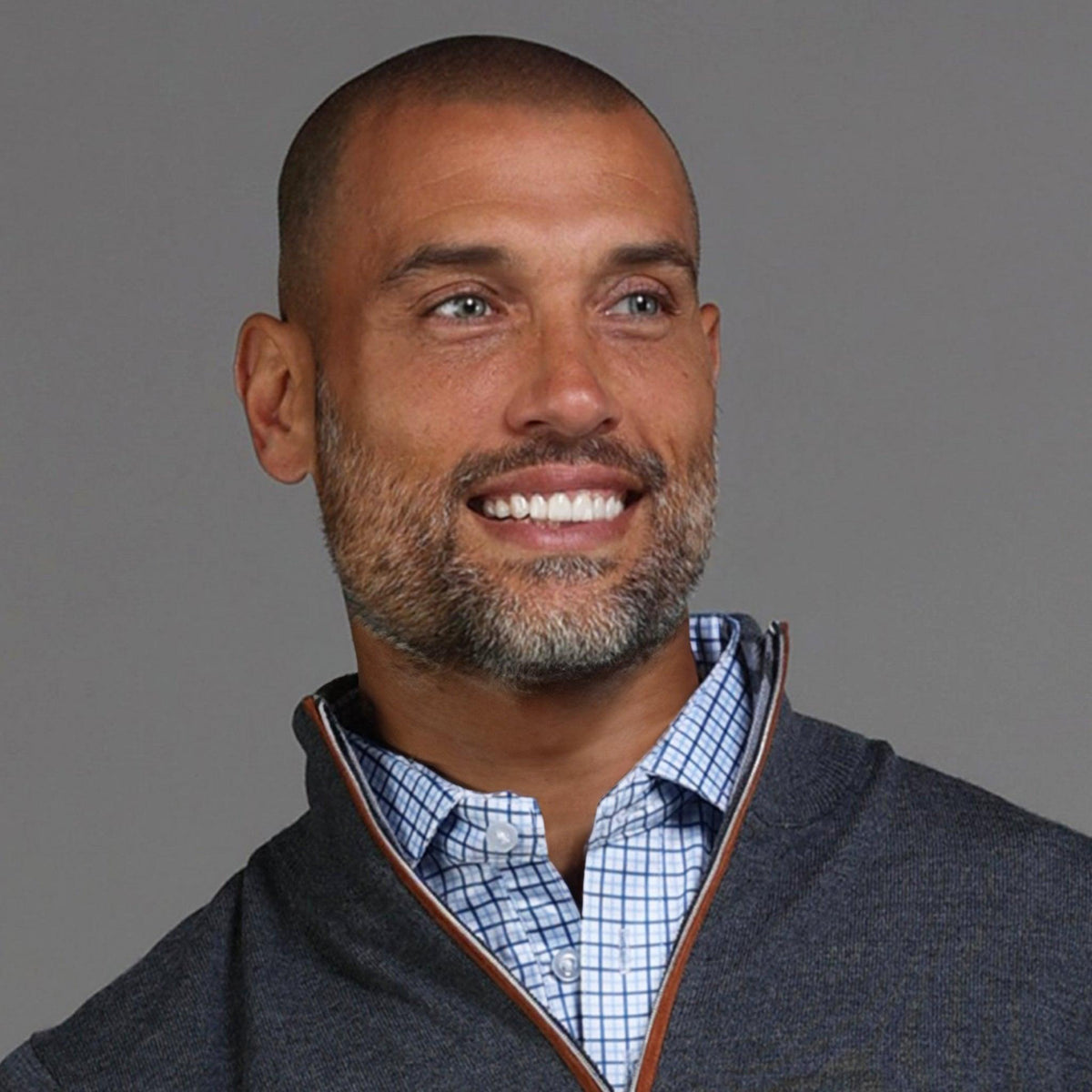 A smiling man with a shaved head and short beard wears the COLLARS AND CO Charcoal with Brown Suede Trim Cashmere Touch Australian Merino Quarter Zip Sweater over a blue checkered shirt, standing against a plain gray background.