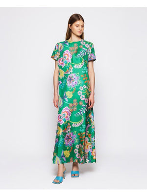 A woman poses elegantly against a white backdrop, her long brown hair flowing over the MIRTO 1956 - FLORAL PRINT SILK DRESS. This green column dress features a Liberty floral print in vibrant pinks, purples, and yellows on silk twill, paired with chic blue open-toe heels.