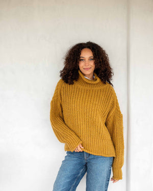 A woman with curly hair stands against a white wall, wearing a chunky mustard-colored MERSEA - BEACHSIDE TURTLENECK and blue jeans. She holds the hem of her turtleneck with one hand and has a relaxed expression.