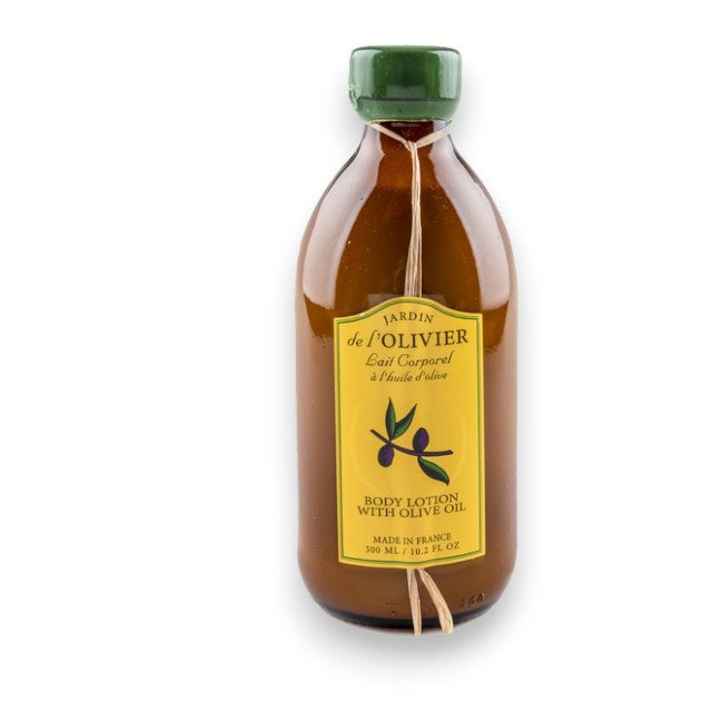 A brown glass bottle with a yellow label featuring an olive branch illustration and the text &quot;BAUDELAIRE - Jardin de l&#39;Olivier Olive Oil Body Lotion with vitamins A, B1, and E.&quot; The bottle is tied with twine at the neck and contains 300 ml (10.2 fl oz) of product. Made in France. Natural scent included.