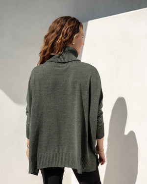 A woman with wavy hair is smiling and looking to the right, wearing a cozy green MERSEA - CATALINA TURTLENECK SWEATER and black pants. She stands against a white wall, with sunlight casting a shadow behind her.