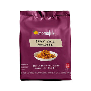 A package of MOMOFUKU Spicy Chili Noodles in burgundy is displayed. The label showcases a bowl of noodles beneath the text "Spicy Chili Noodles." Weighing 16.75 oz (475g), it contains five servings of air-dried noodles mingled with a robustly spicy sauce, delivering a vegan and soy-rich flavor experience.