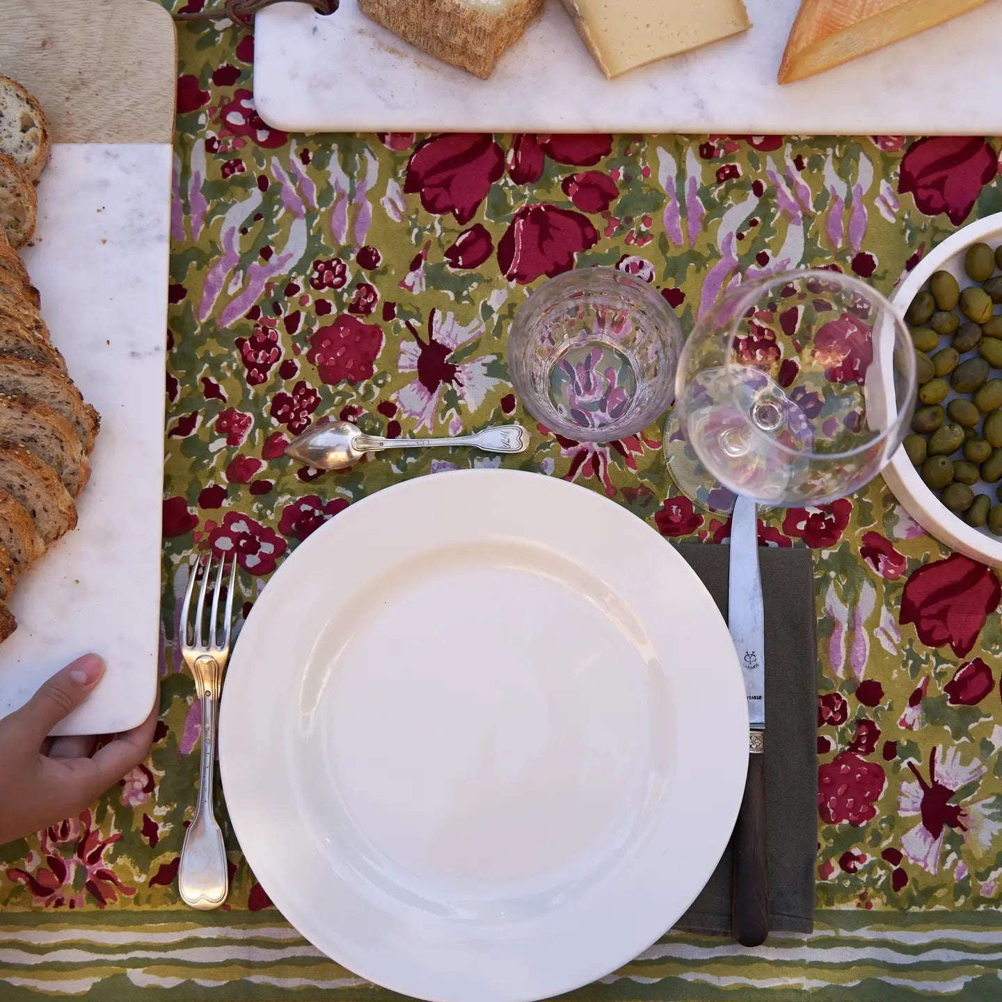 A table setting includes an empty plate, cutlery, two glasses on a COULEUR NATURE JARDIN RED AND GREEN TABLECLOTH 59X59. To the left is sliced bread on a board, a cheese assortment at the top, and a bowl of green olives to the right. A hand reaches for the bread amid Southern France charm.