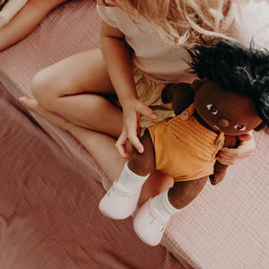 A close-up of a baby wearing OLLI ELLA USA DINKUM DOLL SHOES in Mallow Pink and white socks. The baby is seated on a soft, pale pink textured blanket, dressed in light brown clothing. Only the lower half of the baby's body is visible.