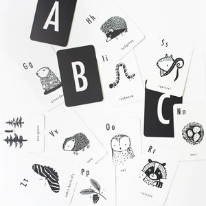 A set of WEE GALLERY - WOODLAND ALPHABET CARDS from Wee Gallery is displayed in an open white box, showcasing high-contrast illustrations of woodland animals and letters. Next to the box on a white surface lies a separate black card holder with the brand name "WEE GALLERY" and a leaf design, enhancing your child's learning experience.