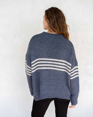 A person with long, wavy brown hair is wearing the MERSEA - SEACOAST STRIPE SWEATER, a loose-fitting, cozy blue knit sweater with white stripes on the sleeves, paired with black pants. They are standing against a plain, light-colored background and facing left, with their hands near their face.