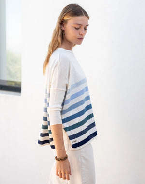 Wearing the MERSEA - CATALINA CREWNECK SWEATER in versatile light pink, a curly-haired individual stands against a plain background. Styled with light-colored pants, hoop earrings, and layered necklaces, they embody the charm of a year-round hero.