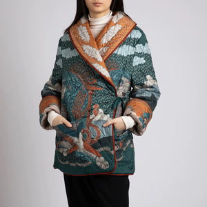 A person wears the SABINA SAVAGE - THE SNOW LION QUILTED JACKET from SABINA SAVAGE ENGLAND, featuring a colorful, intricately designed nature-themed pattern with trees, sky, and animals. Reminiscent of a reversible silk twill jacket, it features a wide collar, contrasting orange inner lining, and large pockets. The person is shown from the shoulders down.