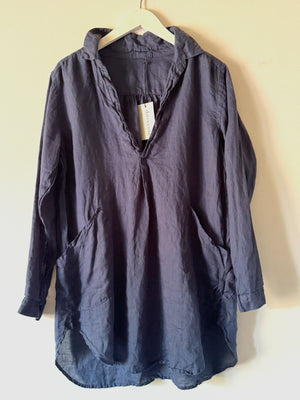 A long-sleeved, dark blue CP SHADES - TETON TUNIC from the brand CP SHADES hangs on a white hanger against a light background. The 100% linen tunic features a collar, V-neckline opening, and large front pockets. A white tag with text is attached to the neckline.