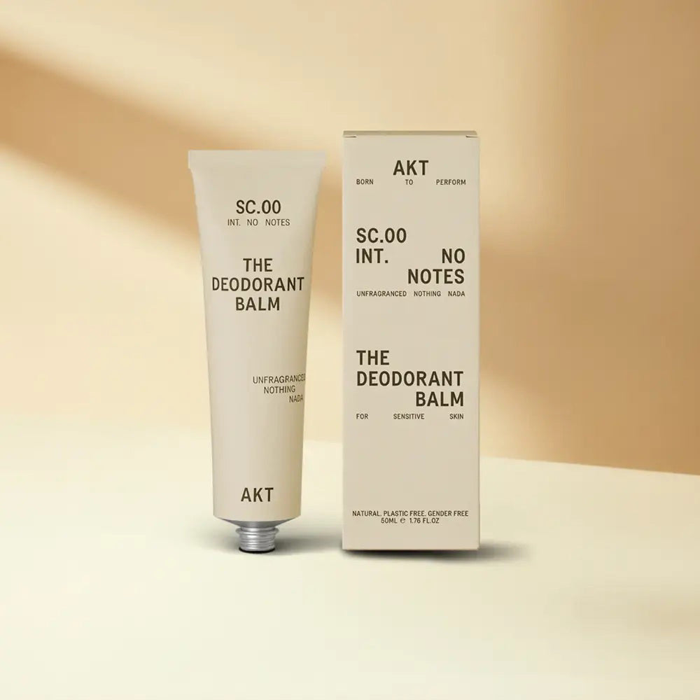 A tube and box of AKT LONDON - DEODERANT BLAM FRAGRANCE FREE, an aluminum-free deodorant, are shown on a beige surface with matching background. The minimalist cream packaging displays black text and the AKT LONDON brand.