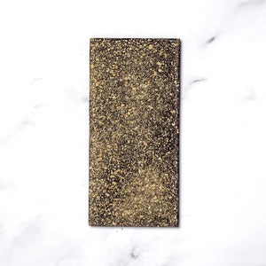 The RITUAL CHOCOLATE - SMORES CHOCOLATE BAR 70% features a rectangular shape adorned with a speckled golden topping, reminiscent of marshmallow flavors, set against a white marble background. The bar has a slightly textured surface due to the glittery gold flecks covering it.