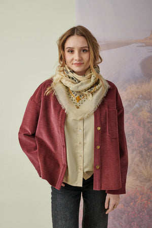 A young woman with light brown hair wears the STORIATIPIC Nelly Square Cashmere and Wool Embroidered Bandana Scarf, paired with a light gray top, standing before a softly colored nature-themed backdrop.