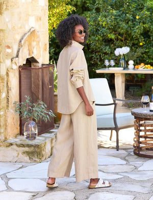 A woman with curly hair, wearing sunglasses, a beige long-sleeve shirt, and a matching pair of FRANK & EILEEN Maisie Wide Leg Linen Pull On Pants from the Tuscany Getaway Set, stands outdoors. She is smiling and leaning against a rustic stone wall. Nearby is a table with a wine bottle and glass, and a white chair under a lush green tree.