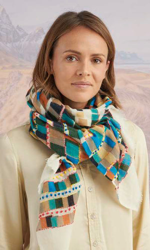 A woman faces the camera, wearing a white button-up shirt, blue jeans, and the STORIATIPIC - ANOUK WOOL SCARF by STORIATIPIC in colorful plaid. The backdrop features soft mountains and grassy plains.