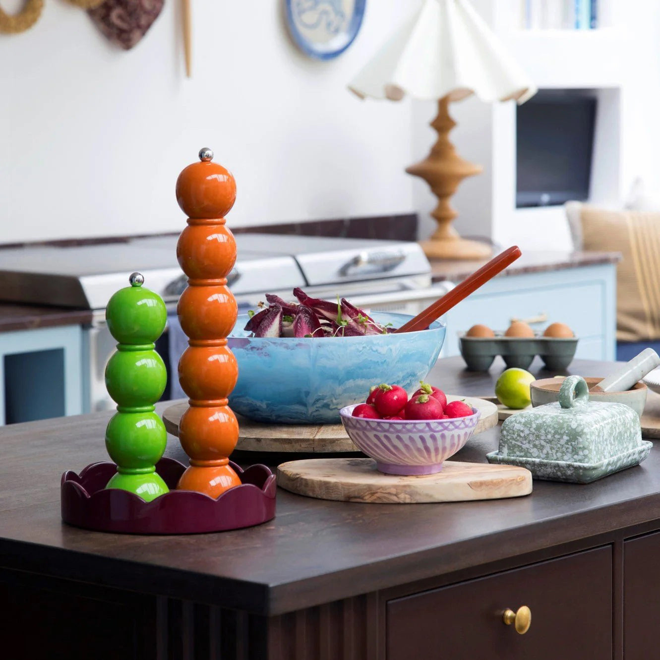 A kitchen countertop featuring a stack of colorful ceramic spheres, a bowl of red onions, a small dish of radishes, a butter dish, and a bowl of eggs. Nearby stands the sleek ADDISON ROSS LONDON - GRANDE SALT OR PEPPER MILL 35CM by ADDISON ROSS. In the background, there is a stove, cabinets, a lamp, and kitchen utensils.