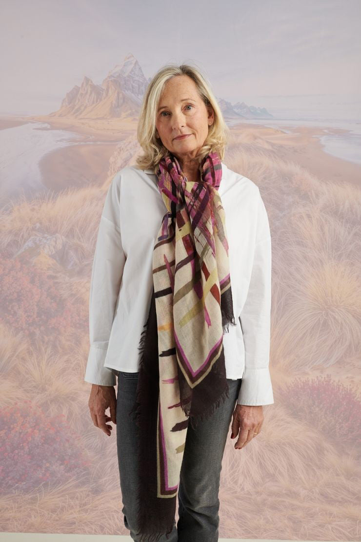 A woman in a blue shirt and jeans stands holding the STORIATIPIC - JULIE WOOL SCARF, a luxury plaid accessory in earthy tones. Behind her, mountains, beach, and grassy dunes create a scenic backdrop.