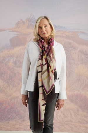 A woman in a blue shirt and jeans stands holding the STORIATIPIC - JULIE WOOL SCARF, a luxury plaid accessory in earthy tones. Behind her, mountains, beach, and grassy dunes create a scenic backdrop.