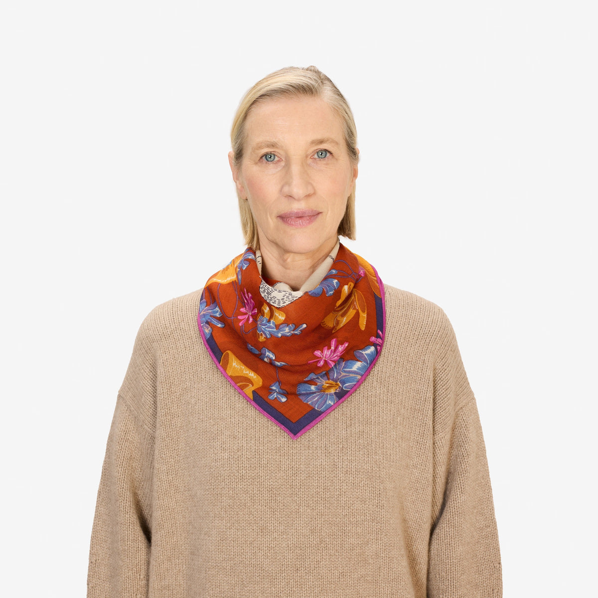A woman with blonde hair, wearing a tan sweater and the INOUI TOUTOU DOG SCARF IN ORANGE, stands facing the camera against a plain white background.