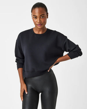 A woman stands confidently against a plain white background, wearing the SPANX Aireessentials Crew Neck Pullover and matching sweatpants made from luxurious comfort spacer fabric. She has her hair pulled back and her left hand is raised slightly. She smiles gently with her lips closed.