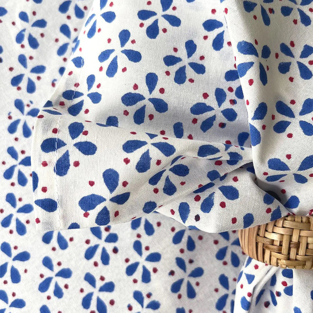 The CHRISTINA DICKSON HOME - PETALS COTTON NAPKIN showcases a blue and red floral pattern on white fabric, crafted in India. Made from 100% cotton, it is knotted over a rattan circle, creating an elegant and textured drape.