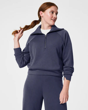 A woman with long brown hair is wearing the SPANX AIRESSENTIALS HALF ZIP sweatshirt and matching pants, both crafted from lightweight fabric for luxurious comfort. She holds her braided ponytail with one hand while looking to her right against a plain white background.