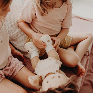 A close-up of a baby wearing OLLI ELLA USA DINKUM DOLL SHOES in Mallow Pink and white socks. The baby is seated on a soft, pale pink textured blanket, dressed in light brown clothing. Only the lower half of the baby's body is visible.