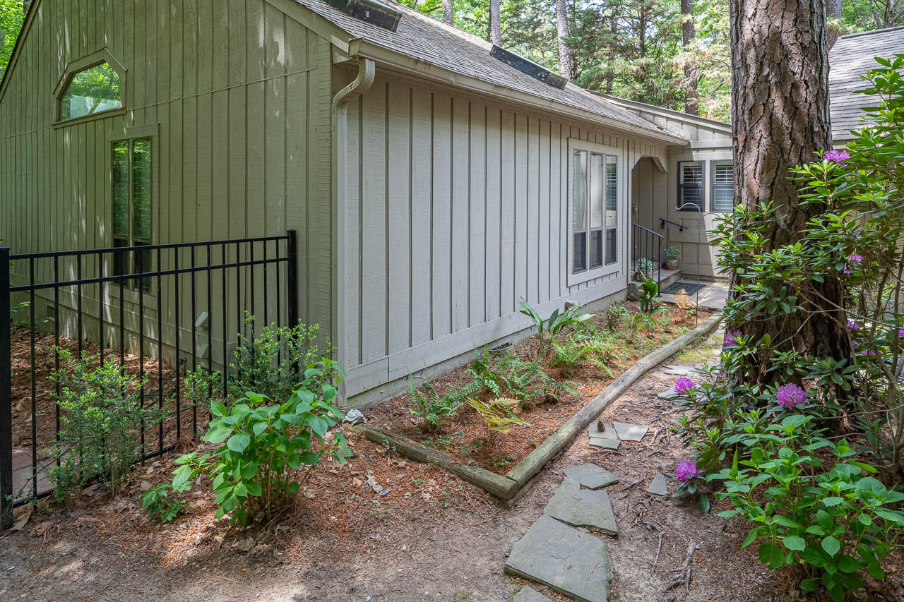 Side view of a light gray wooden house with vertical siding, surrounded by trees and greenery. A narrow garden bed with plants lines the house, and stone pavers lead to a small porch by the entrance.