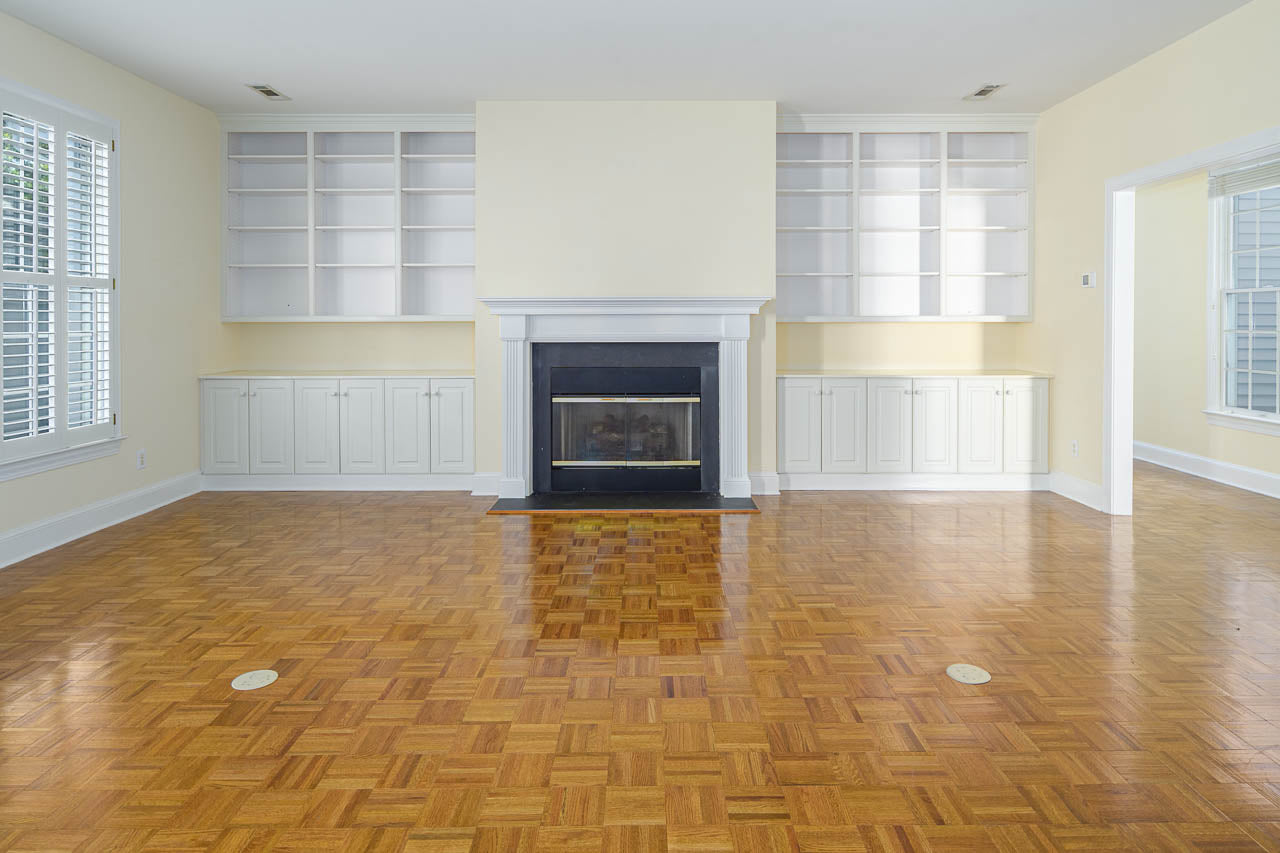 A spacious empty living room with parquet wood flooring, a white fireplace centered between built-in shelves and cabinets, pale yellow walls, and large windows with white shutters.