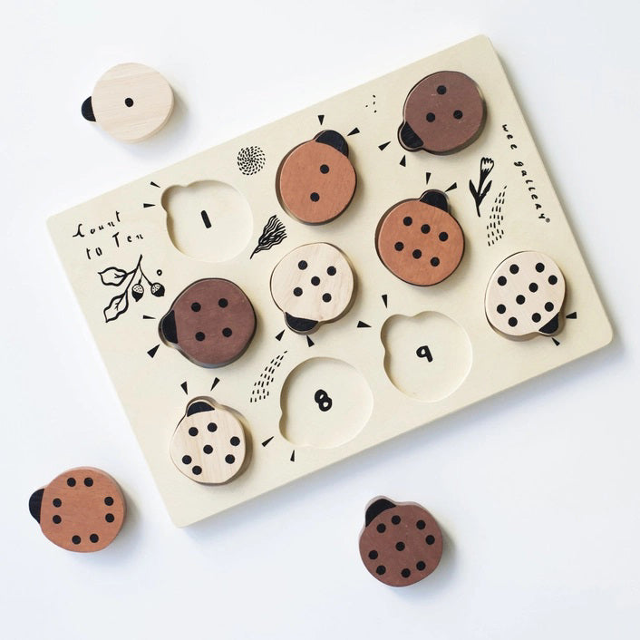 The WEE GALLERY - LADYBUGS COUNT TO 10 TRAY PUZZLE by WEE GALLERY is a wooden puzzle board featuring ladybug-shaped pieces in varying shades from light beige to dark brown. This educational puzzle helps teach counting skills and depicts leaves and twigs alongside the words &quot;Count to Ten&quot; on the left side. Each ladybug is adorned with black dots on their backs to represent number concepts.